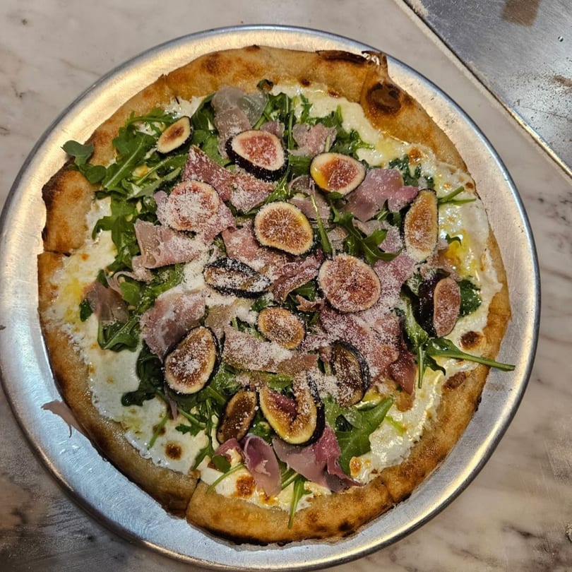 Tino's Delicatessen fig and prosciutto pizza with arugula and parmesan on a metal tray.
