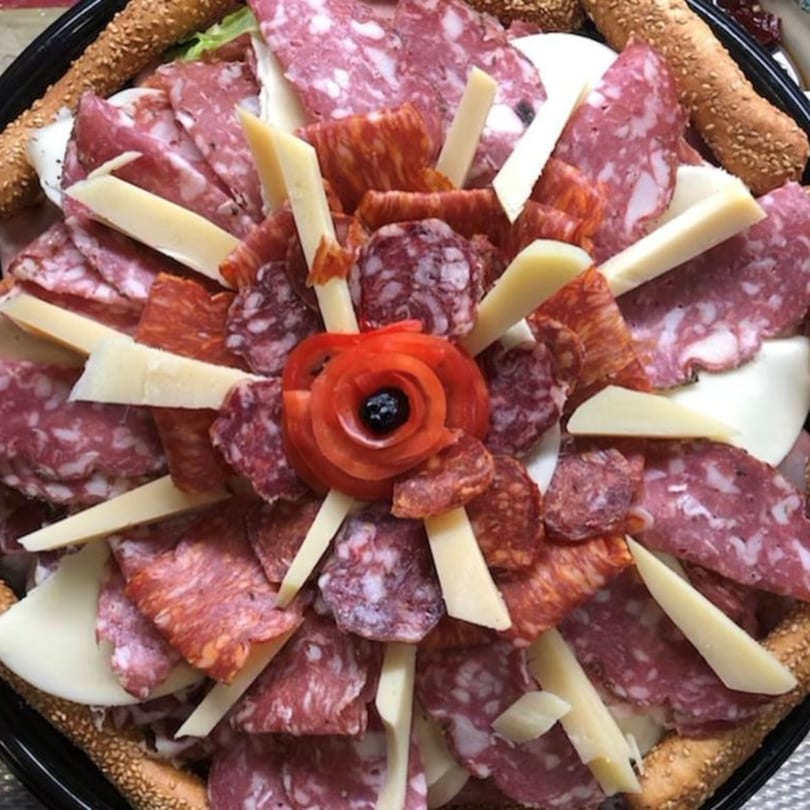 A Tino's Delicatessen charcuterie board with various Italian meats, cheeses, and breadsticks, garnished with a tomato rose.