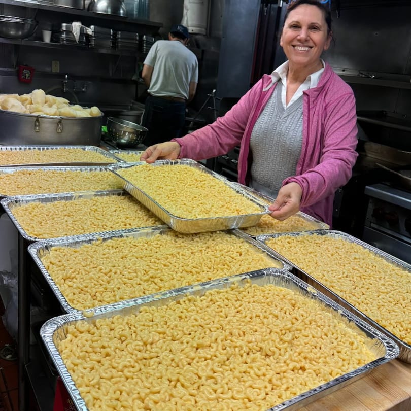 A smiling woman in a commercial kitchen holds a large tray of macaroni and cheese.