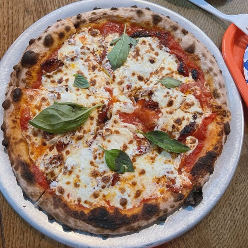 A freshly baked Tino's Delicatessen pizza with melted cheese, tomato sauce, and basil on a metal tray.