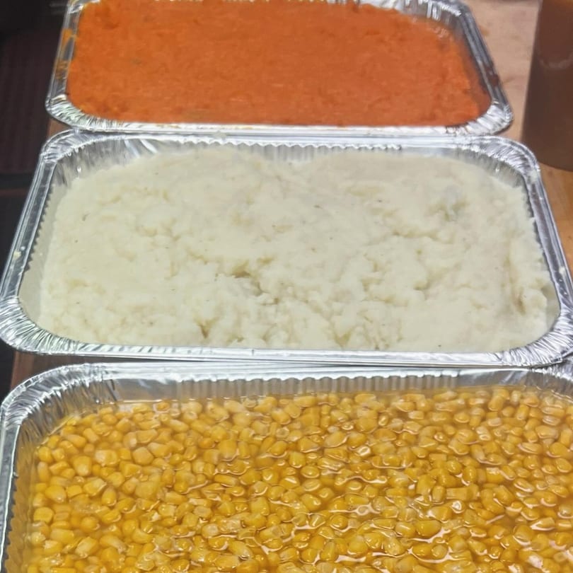 Three foil trays of food: orange mashed sweet potatoes, white mashed potatoes, and yellow corn.