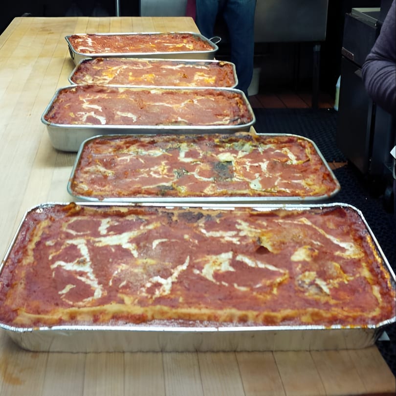 Five large trays of Tino's Delicatessen lasagna with red sauce and cheese on a wooden table.